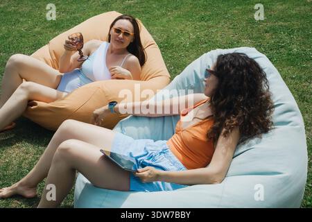 Two cheerful women friends relax on bean bag chairs in a sunny garden drinking and using a digital tablet during summer vacation leisure time. Stock Photo