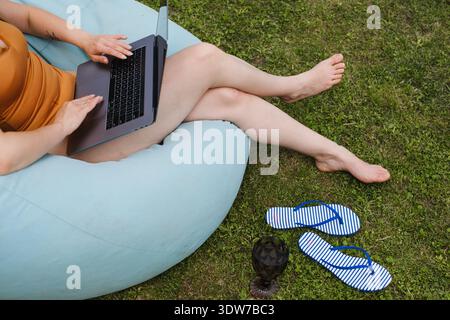 Anonymous woman typing on laptop while sitting on blue beanbag in green garden. Concept of remote work, freelancing and digital nomad lifestyle in Stock Photo