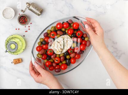A woman holds in her hands a glass baking dish with white  mold cheese (camembert, brie), cherry tomatoes, black and green olives, kalamata, thyme and Stock Photo