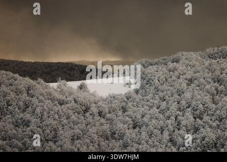 A dramatic winter mountain panorama showing a vast, frost-covered forest under a dark, heavy sky, with a spectacular sunbeam breaking through the clou Stock Photo
