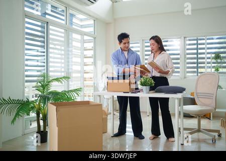 Two diverse adult Asian colleagues planning office relocation. They organize items, discuss notes in a bright modern workspace. Stock Photo