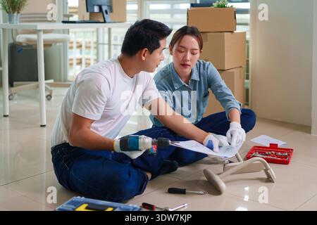 Focused Asian adults collaboratively assemble modern furniture using power drill. They follow manual instructions for a home office setup. Stock Photo
