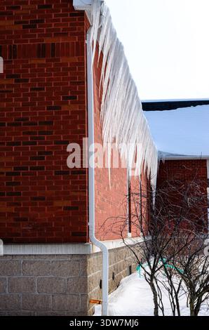 Long row of large icicles hanging alongside a red brick wall on a snowy winter day with a clear hazy sky. Stock Photo