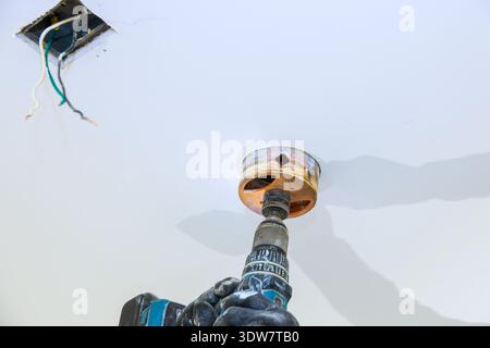 Worker uses power drill to hole ceiling fixture while wires hang above in room. Stock Photo