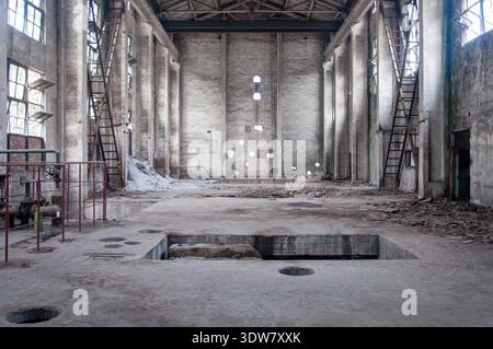 Wide angle interior view of an abandoned factory warehouse featuring high ceilings, large windows, a damaged concrete floor with pits, and scattered d Stock Photo