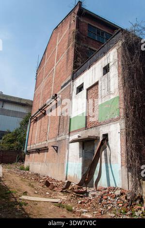 Vertical shot of the side and back of a tall, weathered brick building with patchwork repairs, external pipes, and debris at the base. Stock Photo