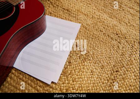 Red acoustic guitar lies on blank sheet music atop a woven straw mat, suggesting songwriting, practice, creativity and relaxed home music-making with Stock Photo