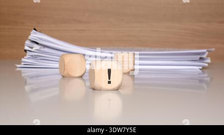 Close up of a wooden block with a printed black exclamation mark, placed on a glossy desk in front of a blurred stack of paper documents. Stock Photo