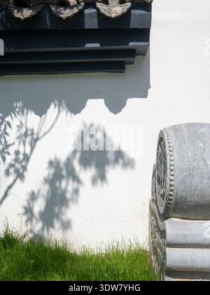 Vertical detail shot of tree branch shadows cast on a white wall of a traditional Chinese building, framed by grey roof tiles and a stone sculpture. Stock Photo