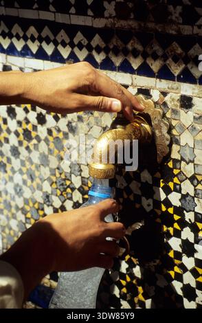 A traveler fills a plastic bottle with fresh water from an engraved brass tap at the historic Nejjarine Fountain, Fes, Morocco. Stock Photo