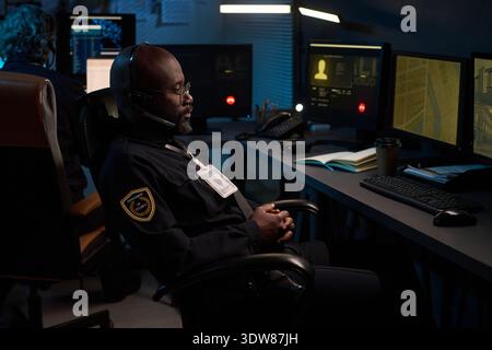 Young Black man sitting at security desk monitoring multiple computer screens with surveillance footage wearing headset and uniform badge, working night shift in control room Stock Photo