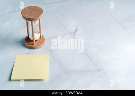 A wooden sand timer next to a yellow adhesive note on a marble surface, perfect for productivity text or reminders Stock Photo