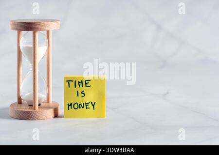 A yellow sticky note with the text "Time is money" placed next to a wooden hourglass on a bright background Stock Photo