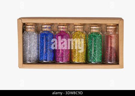 Six small glass bottles filled with different colors of glitter are placed in a cardboard box. Each bottle has a cork top. This setup is ideal for art Stock Photo