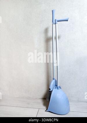 Light blue broom and dustpan set standing against a wall on a tiled floor. Household cleaning tools in minimalistic home interior. Stock Photo