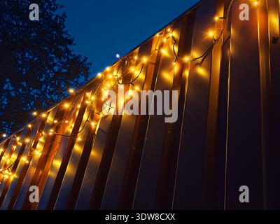 Warm yellow fairy lights hanging on a metal wall at dusk, glowing softly against the deep blue evening sky, creating a cozy festive atmosphere. Stock Photo