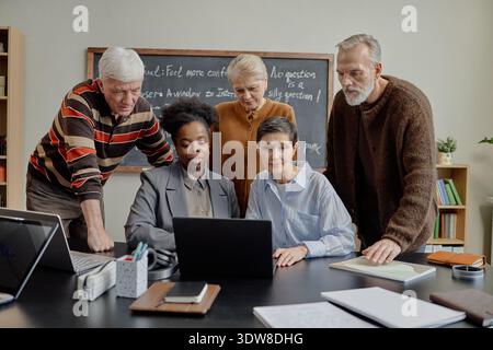 Diverse Group of Senior Adults Collaborating at Laptop in Classroom Stock Photo