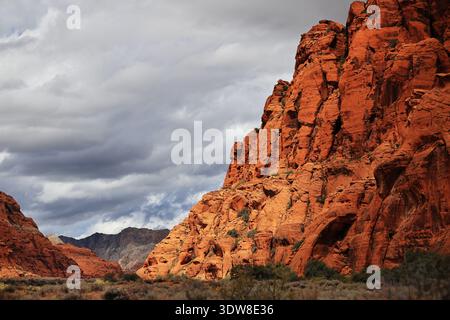 Sunlit steep red sandstone cliff towers above desert shrubs as gray storm clouds sweep across the sky at Snow Canyon State Park near St George, Utah. Stock Photo