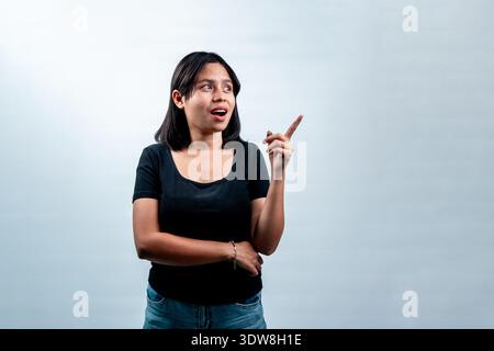 Excited Indonesian woman in black t-shirt pointing aside with open mouth. Promotional announcement and casual youth lifestyle Stock Photo