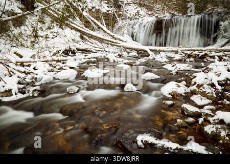 Winter stream with snow-covered rocks and in the background a long exposure frozen waterfall in a snowy woods. Stock Photo