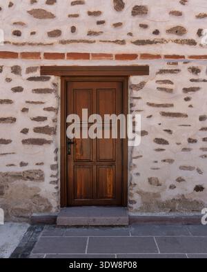 Rustic brown paneled wooden door with black handle in an old white plastered stone wall with exposed stones. Paved walkway in front. Traditional building facade Stock Photo