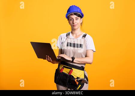 Confident female construction worker stands with laptop in hand, managing project plans digitally. Skilled renovator in hardhat and toolbelt represents modern jobsite coordination and workflow. Stock Photo