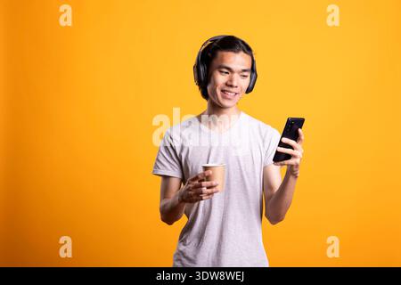 Joyful asian man holding smartphone and coffee in yellow background studio. Happy male individual with headphones enjoys relaxed moment, watching social media videos or texting during casual break. Stock Photo