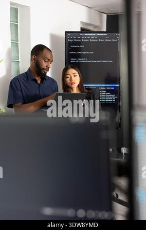 Male and female developers collaborate in tech office, reviewing software data on laptop. Diverse coders analyze HTML scripts together, improving website functionality through teamwork. Stock Photo