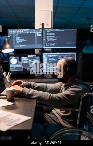 Male programmer wearing headphones works intently at workstation equipped with laptop and multiple displays. The image reflects modern coding routines, concentration, and adaptive technology support. Stock Photo