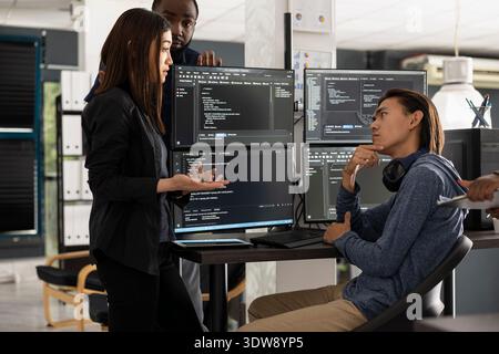 Male and female software developers stand by computer monitors while brainstorming coding project for startup. Diverse team collaborate on refining HTML scripts and building modern website together. Stock Photo