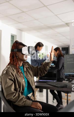 Black female programmer wearing VR glasses interacts with floating 3D interface while working indoors. Futuristic software developer explores augmented reality technology for advanced app development. Stock Photo