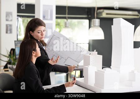 Caucasian manager explains project floor plans to coworker while examining white architectural model. Female design team collaborating in office, planning and ensuring successful construction project. Stock Photo