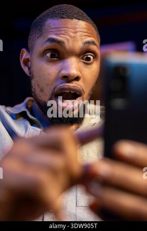 Shocked african american person reading surprising news on his smartphone, sitting in his living room with mouth open. Young adult being surprised while he holds mobile phone. Close up. Stock Photo