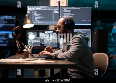 Caucasian software developer works intently on laptop, wearing headphones as monitors glow with technical data. Freelance programmer codes in tech office, reflecting concentration and digital workflow Stock Photo