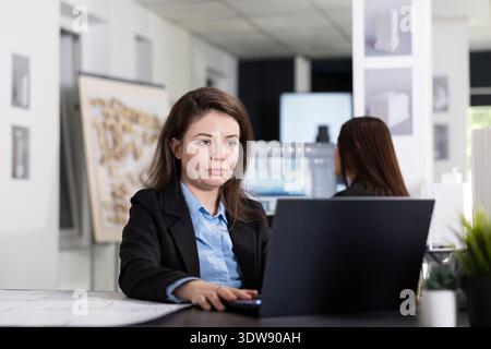 Focused white woman works on laptop, creating reports and planning schedules for upcoming construction site visits. Female architect reviews pending projects and organizes tasks for next inspections. Stock Photo