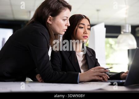 Female colleagues analyze building floor plan on laptop, checking measurements and verifying structural accuracy. Asian and caucasian architects collaborate on design drafts for new construction client Stock Photo