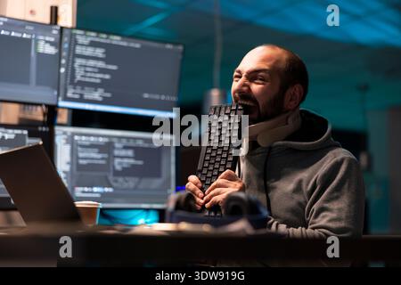 Irritated programmer presses face against keyboard in frustration, overwhelmed by repeated technical glitches. Tired coder stays late in office, angry and stressed as equipment fails to function. Stock Photo