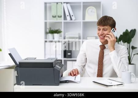 Smiling man taking printed sheet of paper from printer and talking by smartphone at white desk in office Stock Photo