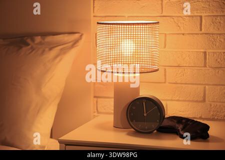 Alarm clock, glowing lamp and sleeping mask on table in dark bedroom, closeup Stock Photo
