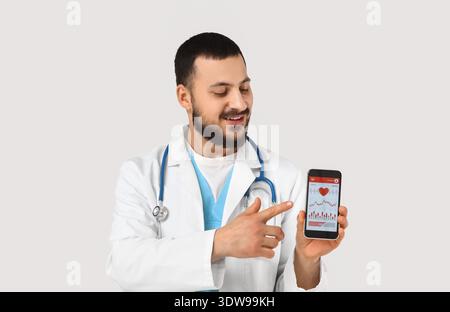 Young happy male doctor with stethoscope pointing at mobile phone on white background. World Health Day Stock Photo