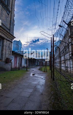 A narrow damp walkway winds between barbed wire fences and brick walls, leading into the yard. A tall razor wire wall stands as glass high rises loom Stock Photo