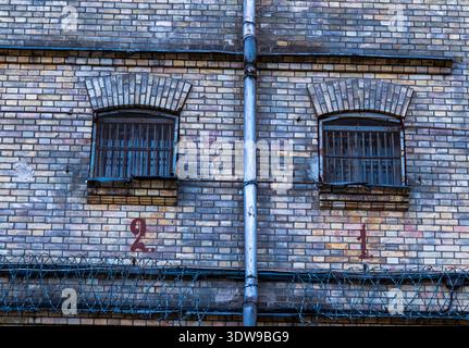 Two small barred arched windows and a rusty drainpipe sit on a brick wall at Lukiskes Prison in Vilnius, Lithuania, with red numerals and coiled barbe Stock Photo