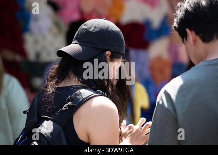 BANGKOK, THAILAND. JAN 25 2026: Woman in black cap and backpack using smartphone in local market, nonverbal communication, side profile Stock Photo