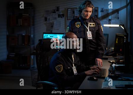 Young Black man wearing headset sitting at desk working on computer, middle aged Caucasian woman in uniform standing beside him observing monitors in security control room Stock Photo