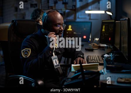 Young Black man wearing headset sitting at desk talking on phone while monitoring computer screens in dark office, visible security badge hanging from neck, working night shift Stock Photo