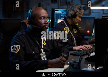 Black man wearing headset monitoring security footage on computer, Caucasian middle aged woman typing on keyboard in control room with multiple monitors and security badges visible Stock Photo