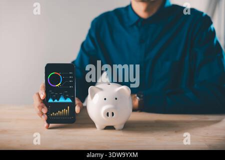 Person holds smartphone displaying digital savings mobile payment app data graphs, beside white piggy bank, representing online financial management, Stock Photo