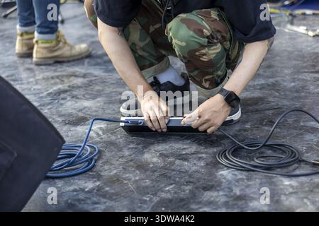 Focused man in camouflage pants working on floor, his hands plugging black cable into power strip during technical setup for an event or production Stock Photo