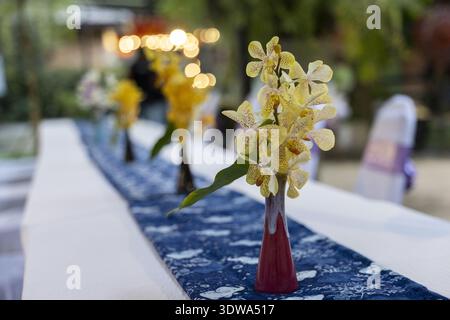 Elegant yellow orchid flower in small red vase on long dining table. beautiful table decoration for romantic wedding reception or banquet with peacefu Stock Photo