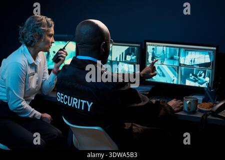 Middle aged Caucasian woman and Black man monitoring surveillance footage on computer screens, discussing security footage and communicating using walkie talkie in control room Stock Photo